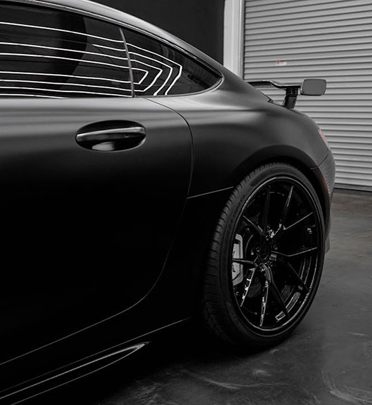 A sleek, matte black sports car with a large rear spoiler and black alloy wheels, showcased against a modern garage backdrop.